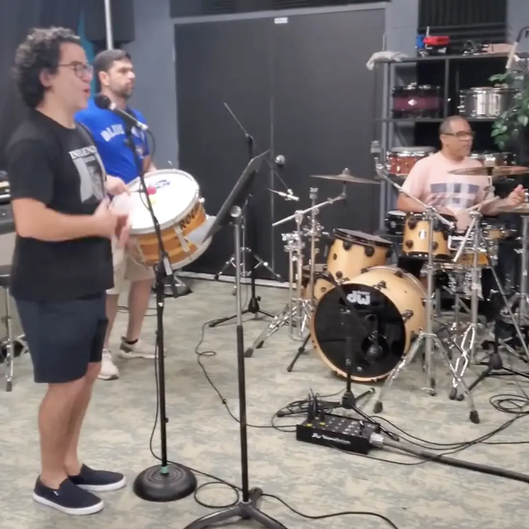 Three musicians playing percussion instruments in a recording studio. A drummer, a percussionist with a surdo, and a singer are visible.