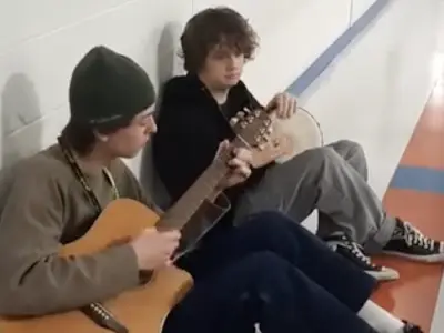 Two teens playing acoustic guitars in a hallway.