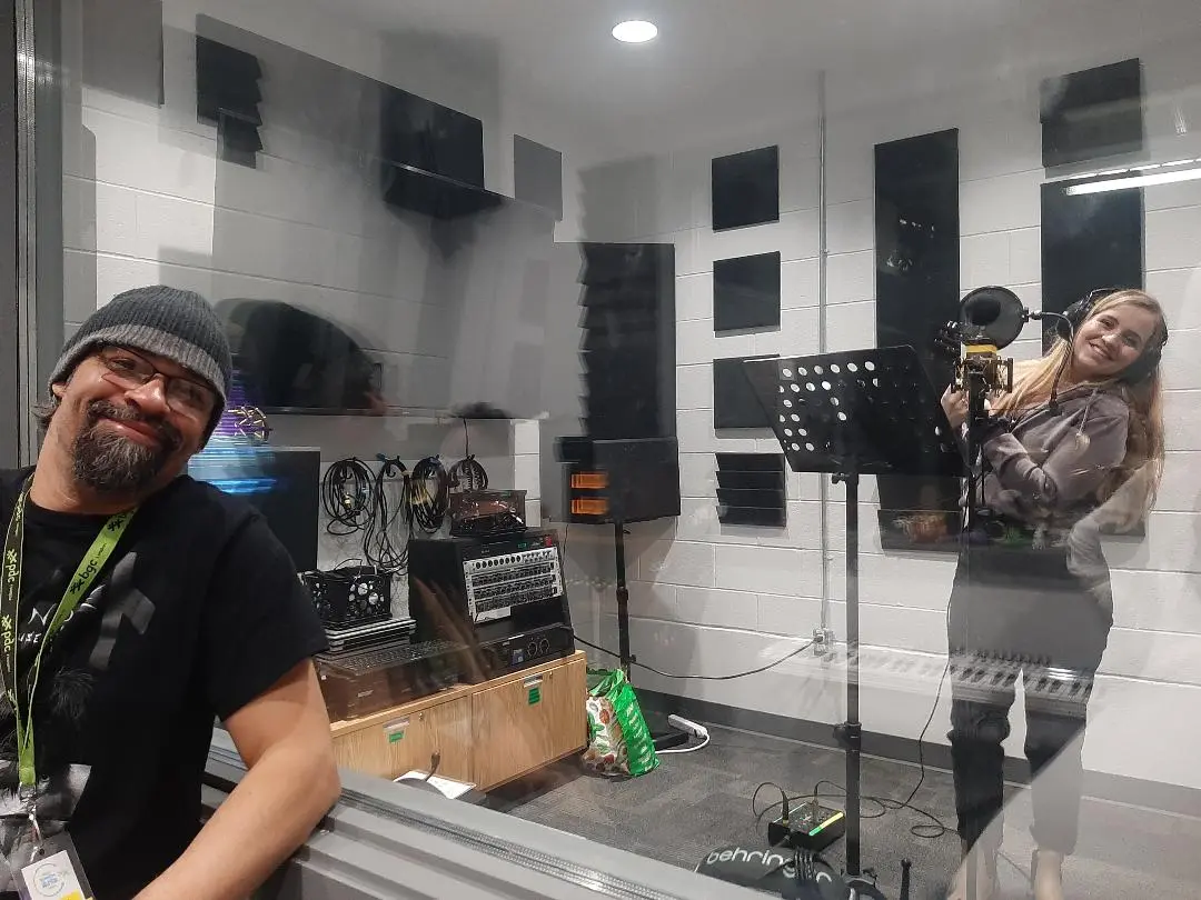Two people in a DCAC Recording Studio; one smiling at the camera, the other recording vocals.